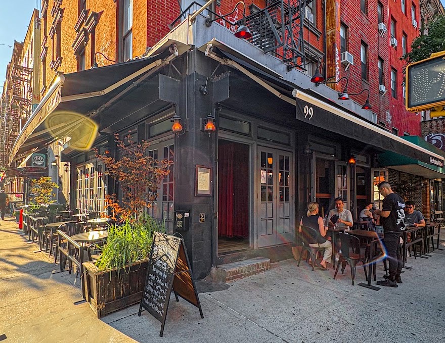 The corner facade of Headless Widow at 99 1st Avenue in the East Village, with black awnings and sidewalk tables
