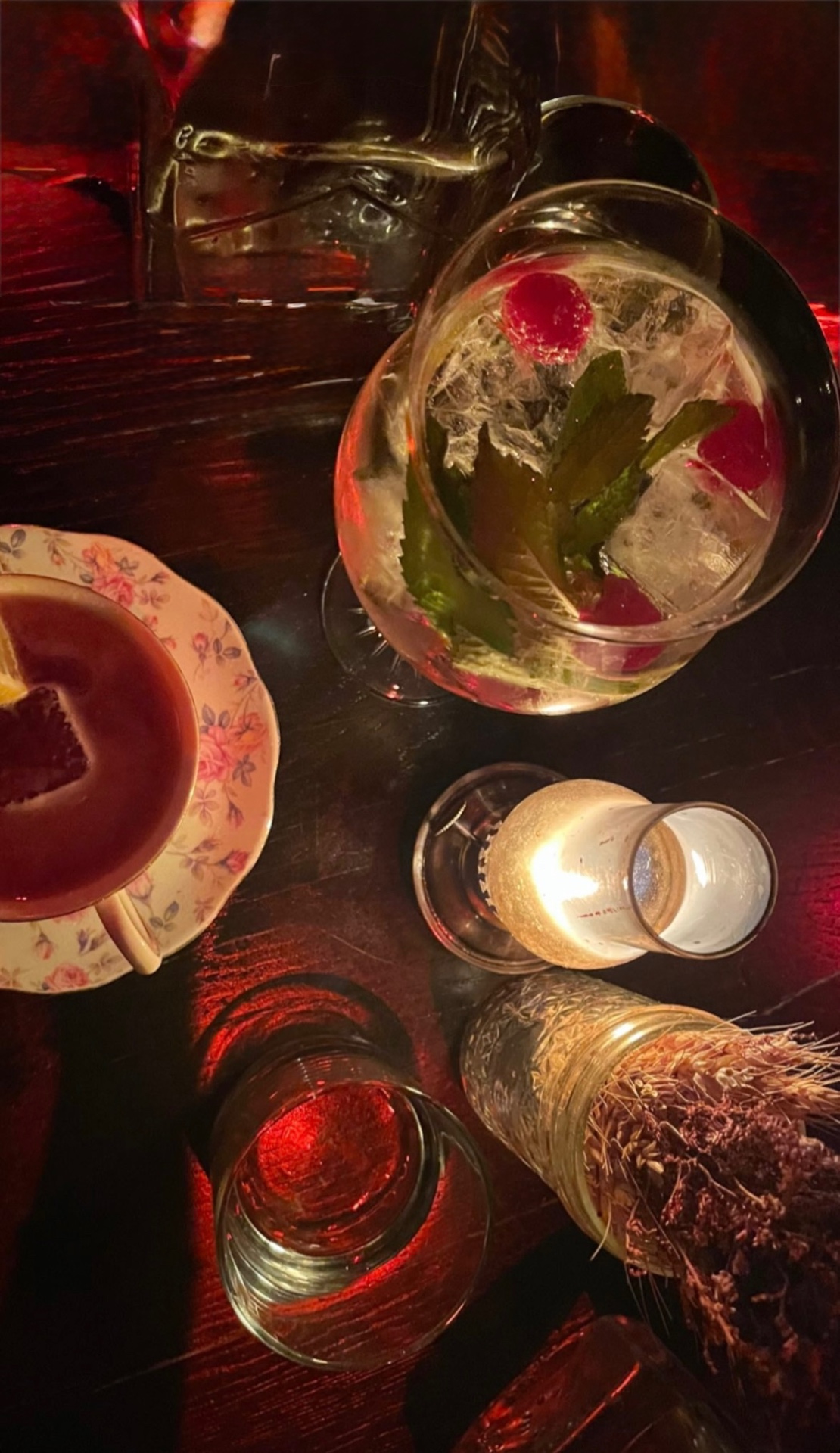 Cocktails and a teacup pour on a dark wood table lit red at Headless Widow