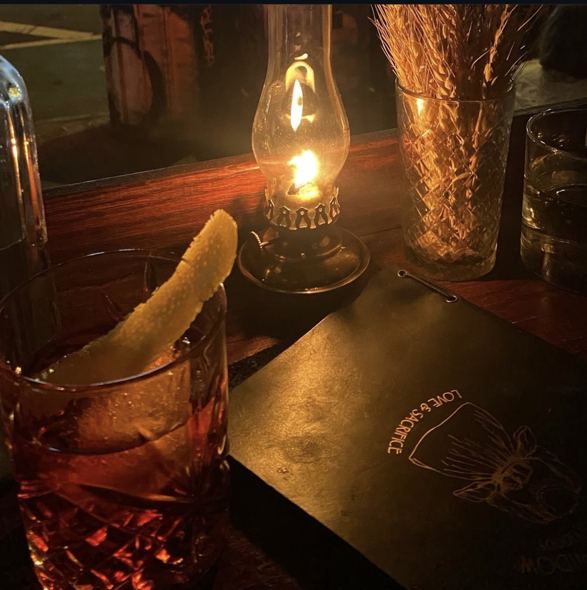 A negroni with a lemon twist beside a lit oil lamp and the Love & Sacrifice menu at Headless Widow