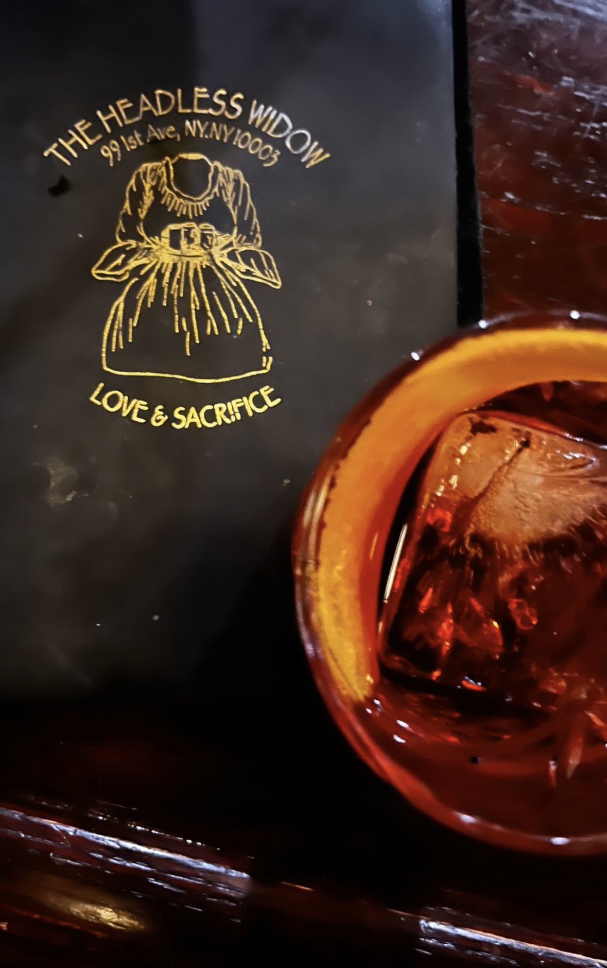 A negroni over a single large ice cube beside the gold-embossed Headless Widow menu reading Love & Sacrifice